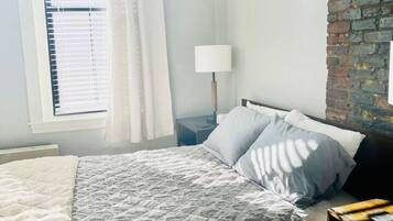 Un dormitorio con cama, mesita de noche, lámpara, ventana con persianas y pared de ladrillo.