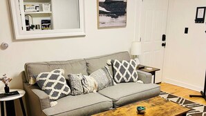 A living room with a sofa, a coffee table, a chair, a small side table, and a wall-mounted shelf. - Cozy Two Bedroom on Munjoy Hill (Portland)