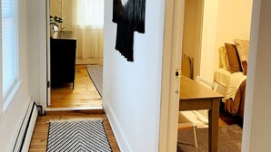 A hallway with a wooden floor, a black and white rug, a door leading to a room with a bed and a chair, and a wall with a black and white abstract art piece. - Cozy Two Bedroom on Munjoy Hill (Portland)
