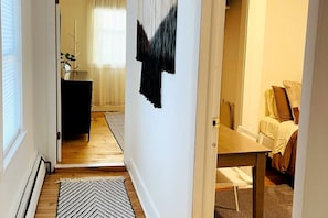 Ein Korridor mit Holzboden, einem schwarz-weißen Teppich, einer Tür zu einem Zimmer mit Bett und Stuhl sowie einer Wand mit einem abstrakten Kunstwerk in Schwarz-Weiß.