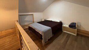 Quarto individual pequeno, com teto inclinado, piso de madeira, cama com colcha branca e escura, mesa de cabeceira e prateleira pequena com um objeto decorativo.