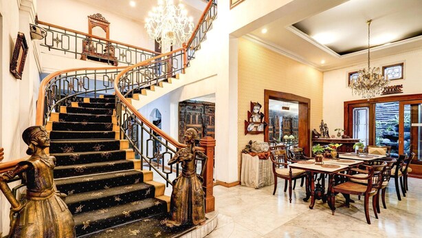 Una imponente escalera con barandillas ornamentales y un candelabro, que conduce a un comedor con muebles de madera y vistas al exterior.