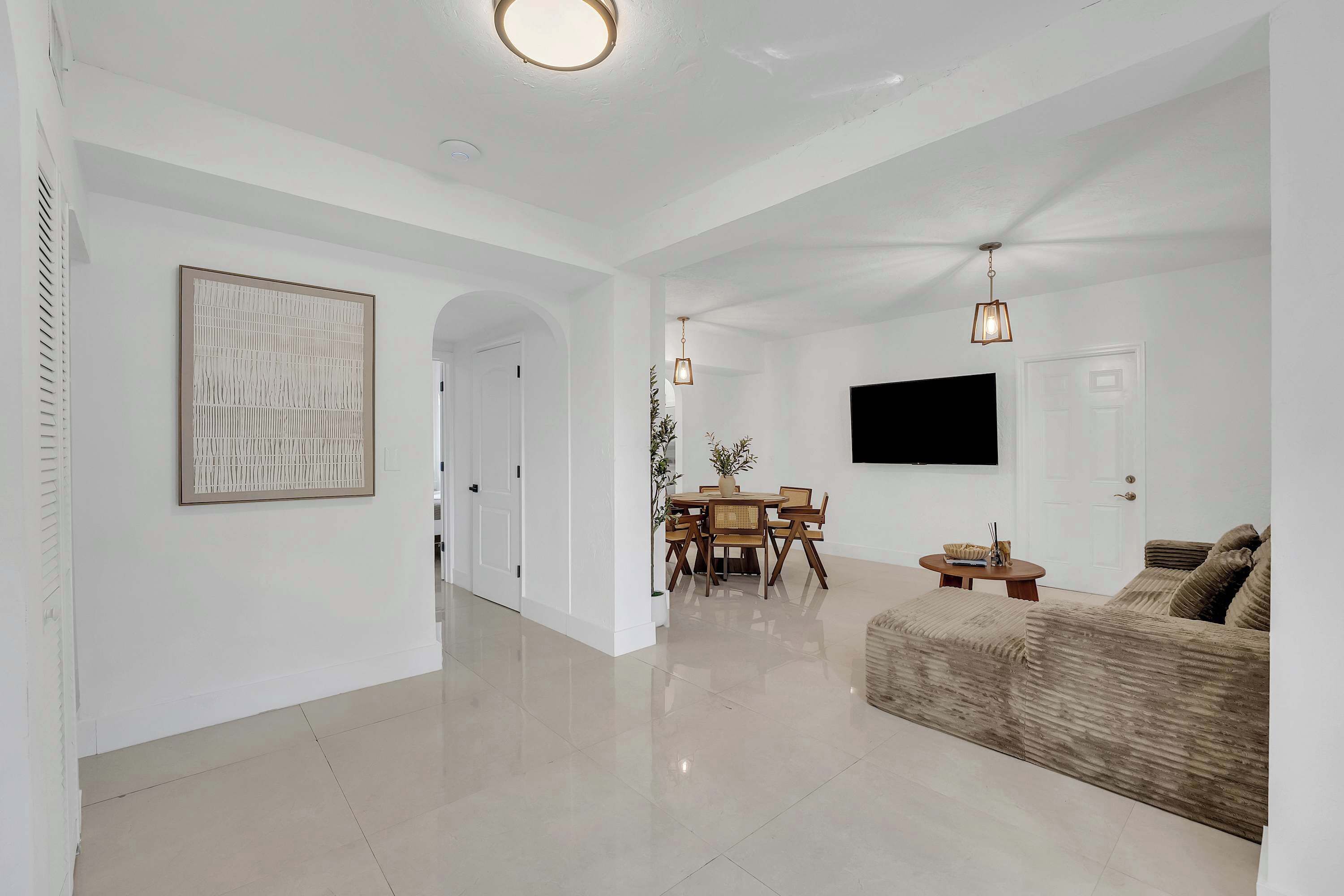 A spacious living area with a sofa, a dining table with chairs, a flat-screen TV mounted on the wall, and a ceiling light fixture.