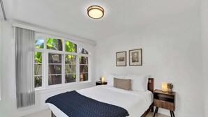 Een slaapkamer met een groot bed, een nachtkastje met een lamp, een raam met gordijnen en lijstjes met foto's aan de muur.