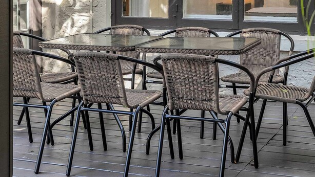 Een overdekt terras met rieten stoelen en tafels, grote ramen en lichtjes.