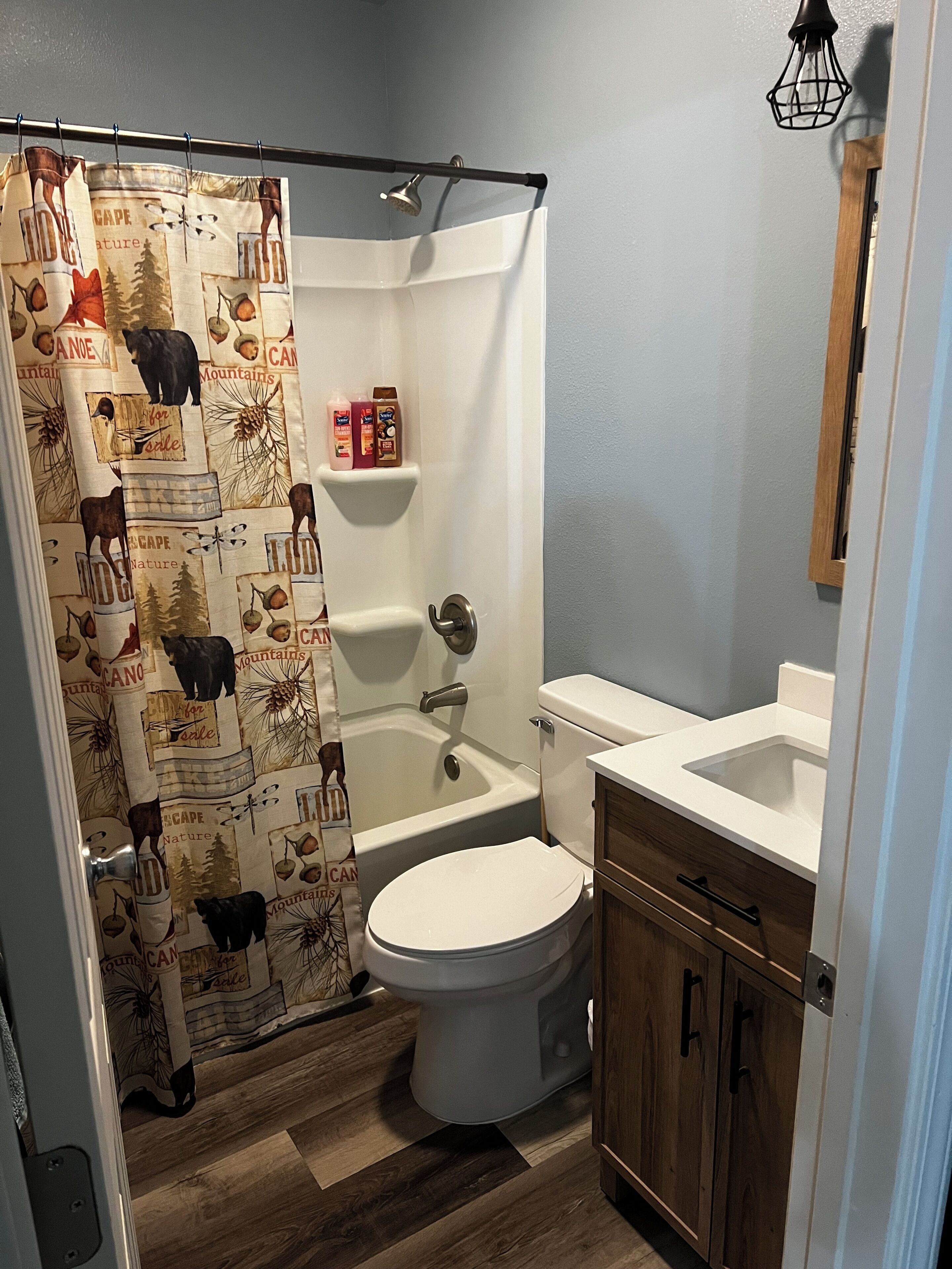 Une salle de bain avec un rideau de douche orné d’un motif représentant la nature, une baignoire blanche, des toilettes et des meubles en bois.