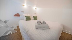 Una cama bien hecha con toallas enrolladas, una silla blanca y una mesita de noche en una habitación sencilla con paredes blancas y una lámpara de techo.