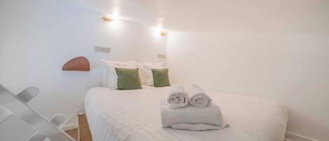 A neatly made bed with rolled towels, a white chair, and a small table in a simple room with white walls and a ceiling light.