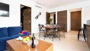 Een moderne woonkamer met een blauwe bank, een houten salontafel met wijnglazen en appels, een eethoek met een houten tafel en stoelen, en een keuken met een barstoel.