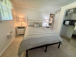 Un dormitorio con cama, dos mesitas de noche, un armario y una puerta que da a otra habitación.