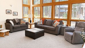 A spacious living room with large windows, a gray sofa set, a coffee table, and a wooden side table with a lamp. - Black Butte Ranch Retreat • 3BR+loft & AC • Walk to Glaze Meadow (Sisters)