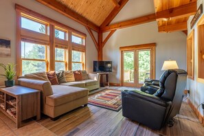 A spacious living room with a wooden ceiling, large windows, a sofa, a massage chair, a TV, and a small table with a plant.
