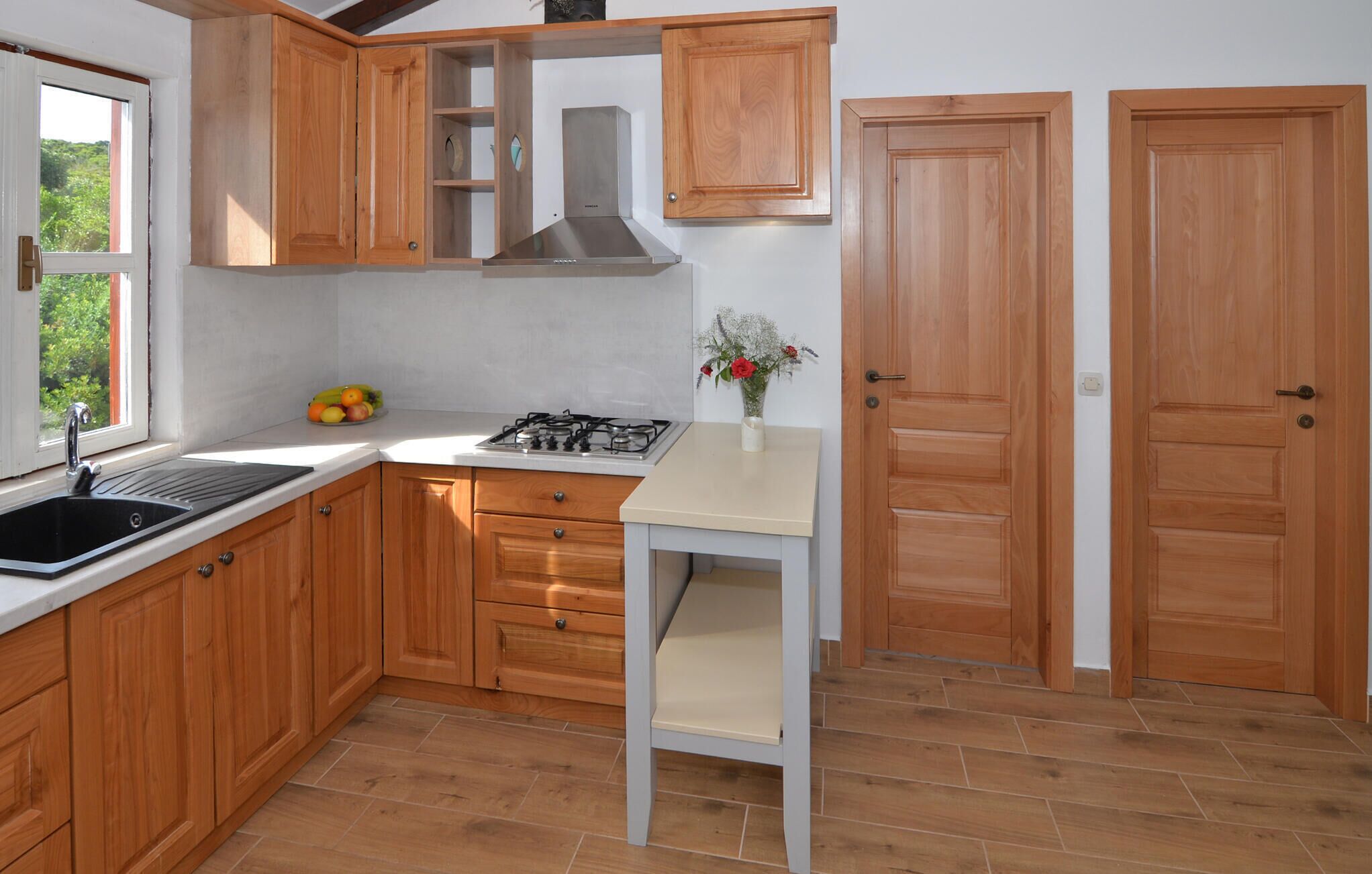 Een keuken met houten kastjes, een gasfornuis, een spoelbak en een raam met uitzicht op groen.