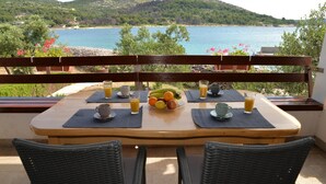 Et spisebord med en skål frugt, to kopper og to glas appelsinjuice, placeret på en balkon med udsigt til havet og bakker.