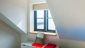 Ein Zimmer mit schrägem Dach, einem Schreibtisch mit Laptop und rotem Notizbuch, einem Fenster mit Blick auf einen Steg und einem kleinen Regal über dem Schreibtisch.
