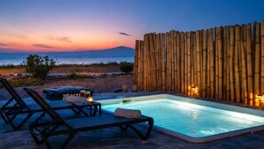 A pool area with lounge chairs and a bamboo wall, overlooking a beach at dusk. - Villa Zeta (Paros)