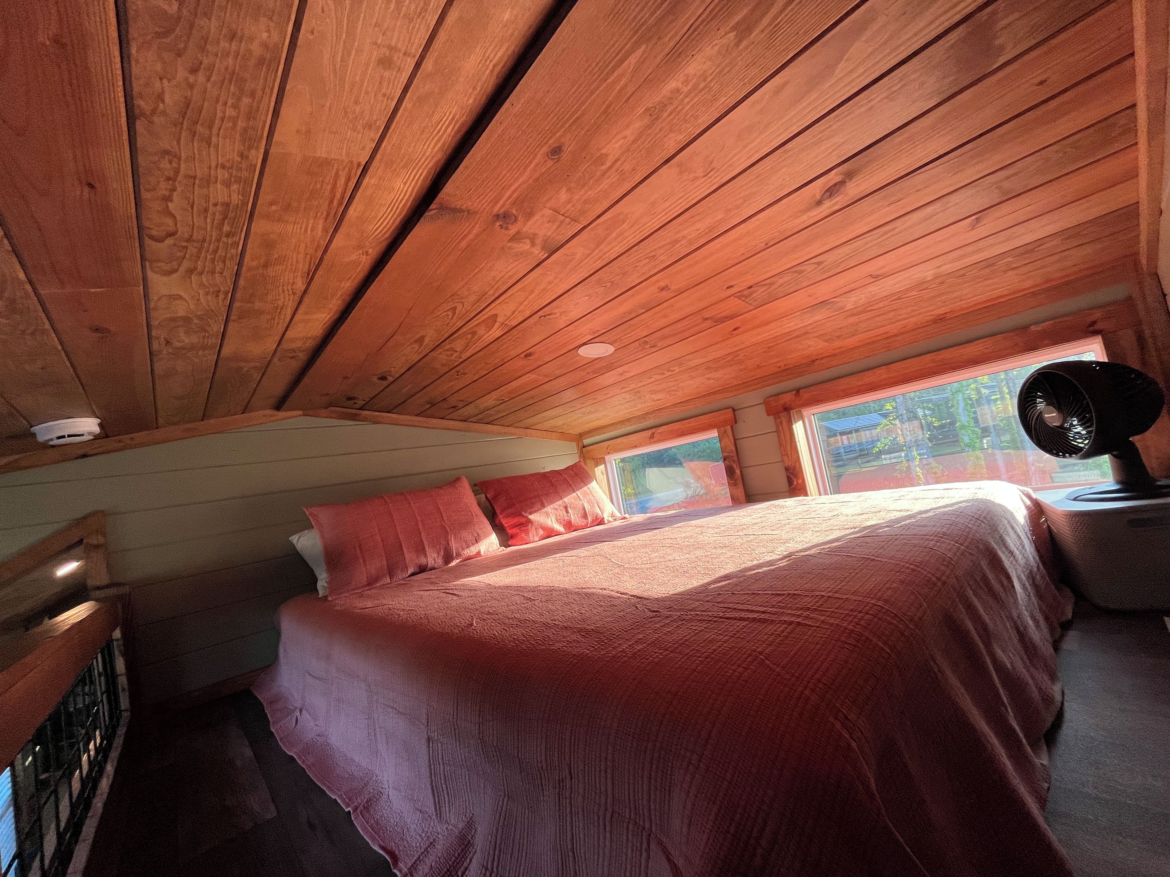 A bedroom with a wooden ceiling, a bed with red bedding, a nightstand, and a window with a view of greenery.