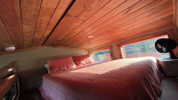 A bedroom with a wooden ceiling, a bed with red bedding, a nightstand, and a window with a view of greenery.