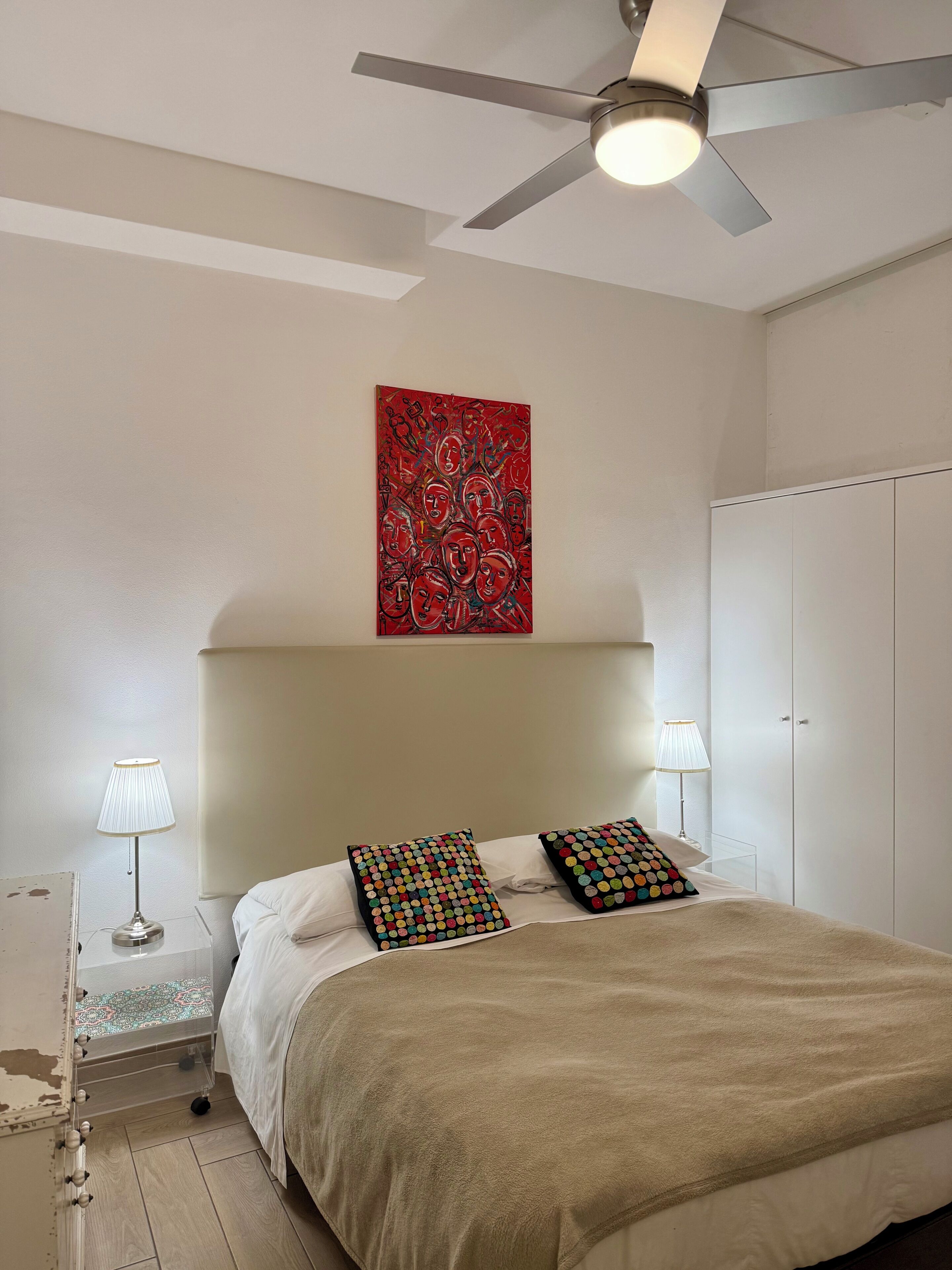 A bedroom with a bed, two bedside lamps, a ceiling fan, a red artwork, and a wardrobe.