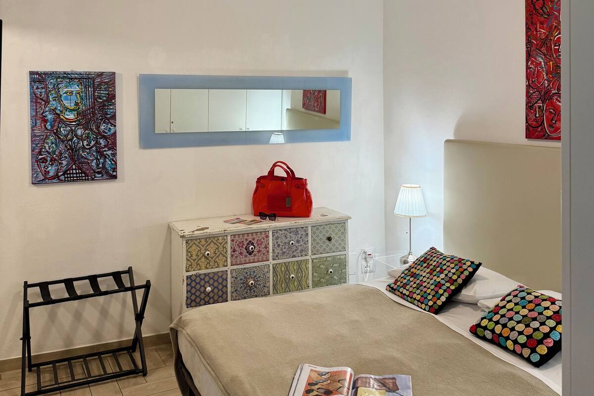 A bedroom with a bed, a ceiling fan, a dresser, a red bag, a mirror, and artwork on the walls.
