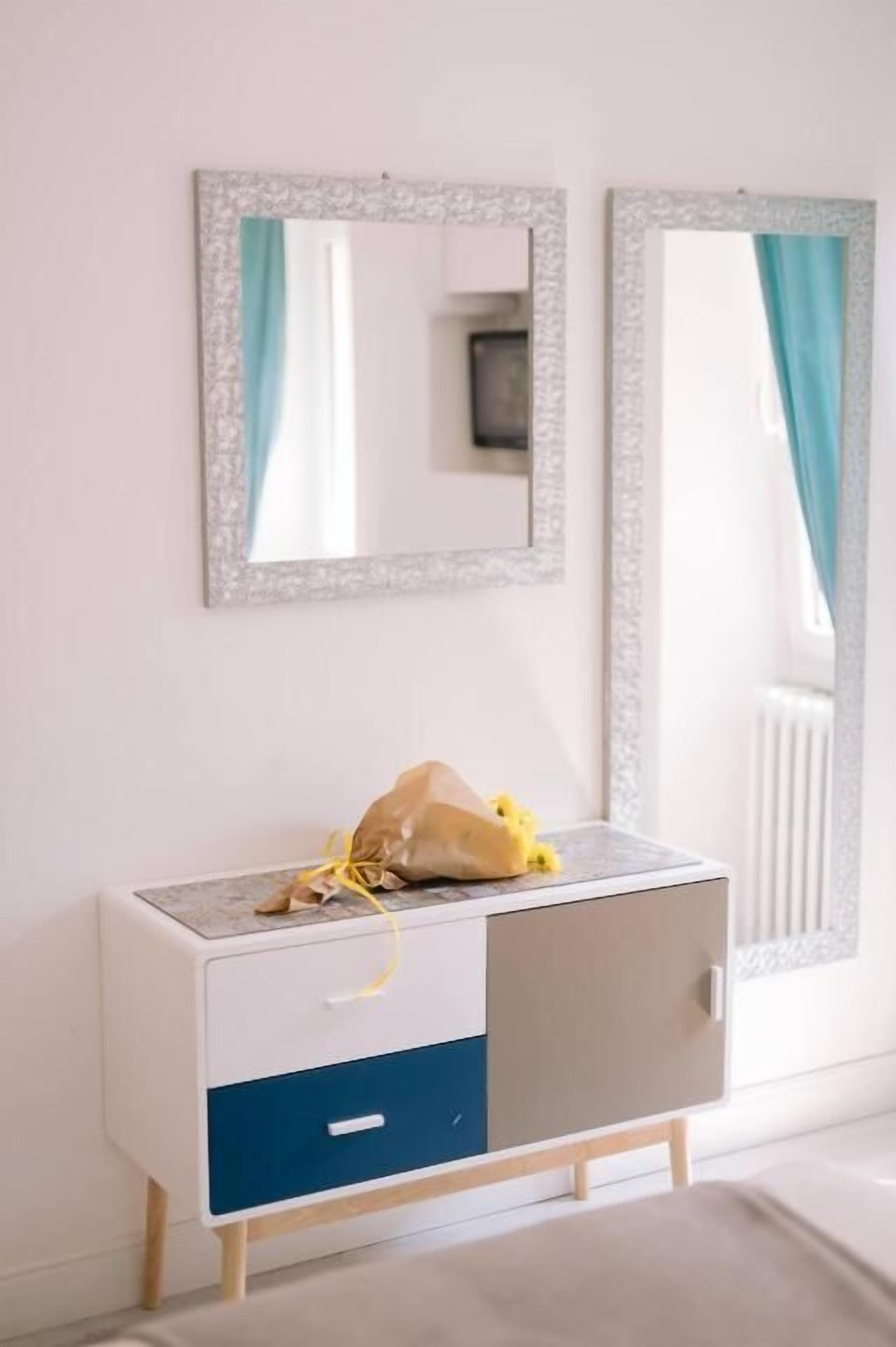 A white and blue cabinet with a mirror above it, a yellow bag on top, and a window with curtains.