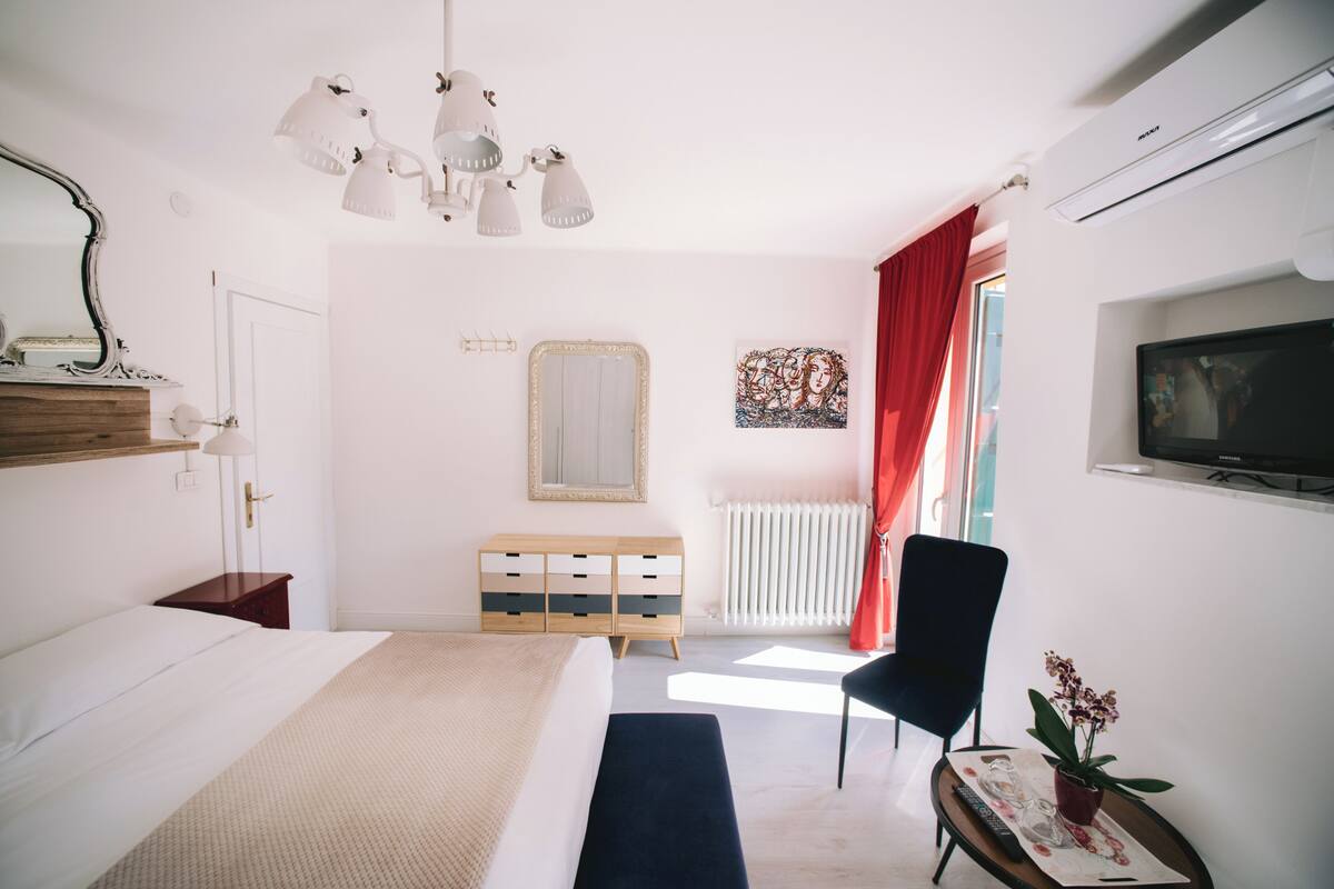 A bedroom with a bed, dresser, mirror, lamp, TV, and a window with curtains.