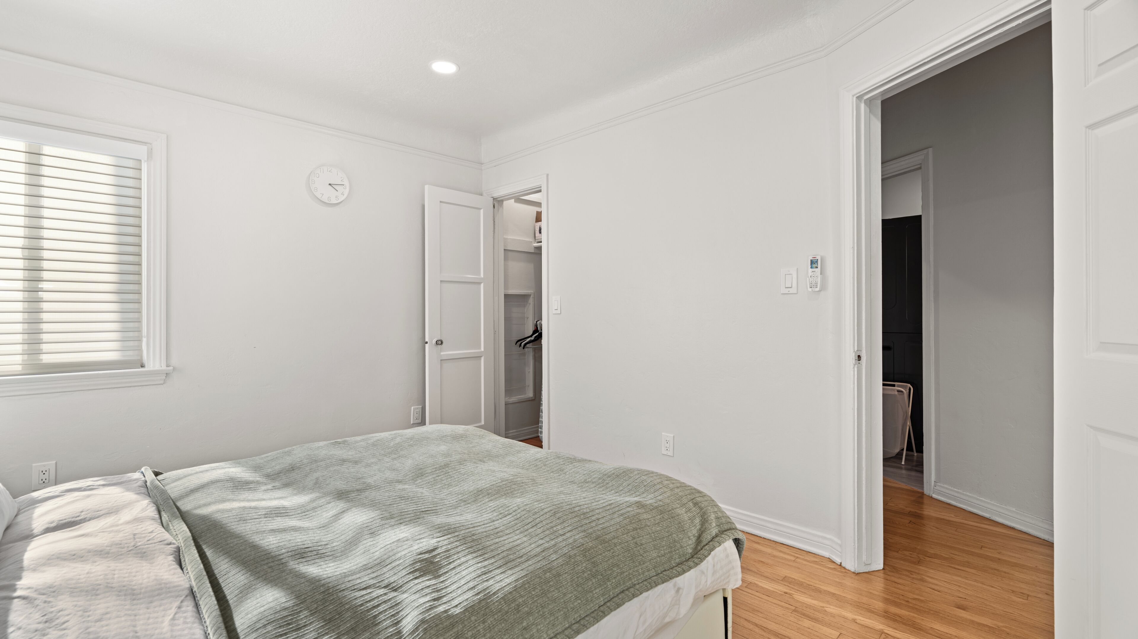 Un dormitorio con cama, una ventana con persianas, un reloj y un armario abierto que da a otra habitación.