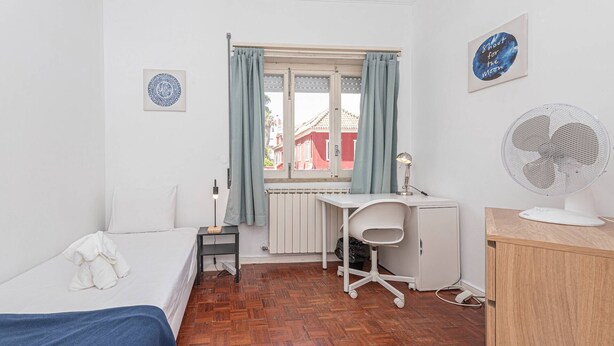 Ein kleines, ordentliches Zimmer mit Bett, Schreibtisch, Stuhl, Heizkörper und einem fenster mit Vorhängen.