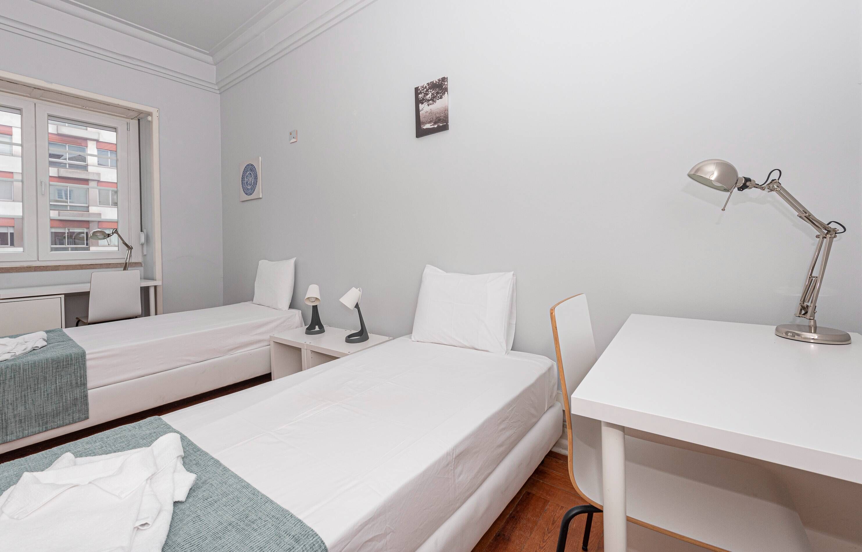 A room with two beds, a desk, a chair, a window, and a wall-mounted picture.