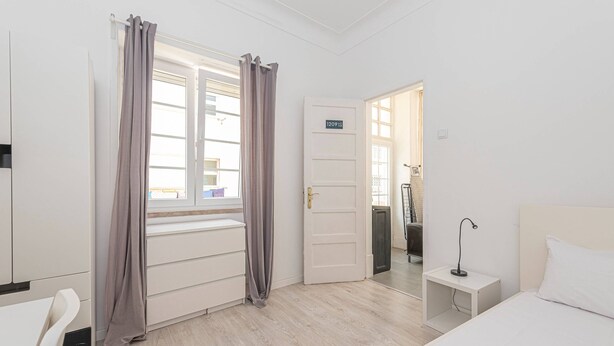 Habitación de hotel con cama, escritorio, silla, ventana con cortinas, puerta con número y mesita de noche con lámpara.