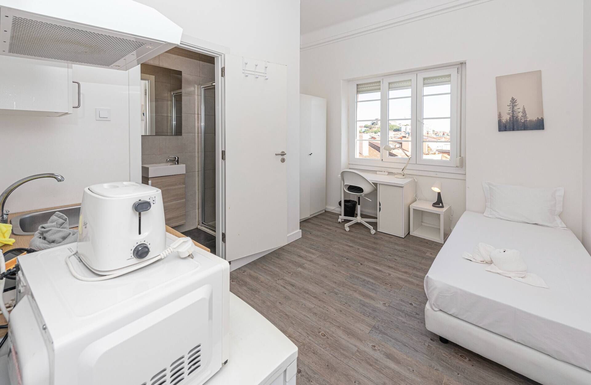 A compact hotel room with a kitchenette, a shower, a desk with a chair, a bed, and a window with a view.