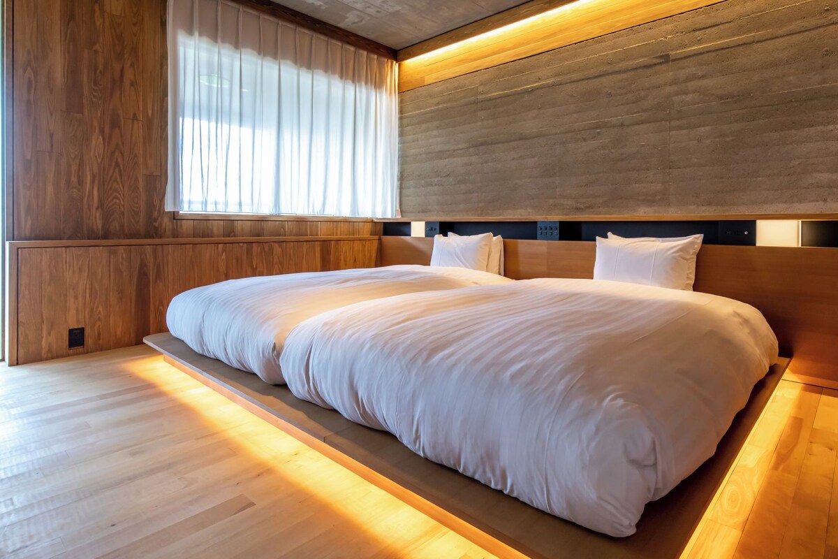 A modern hotel room with a large bed, wooden panel walls, and a large window with curtains.