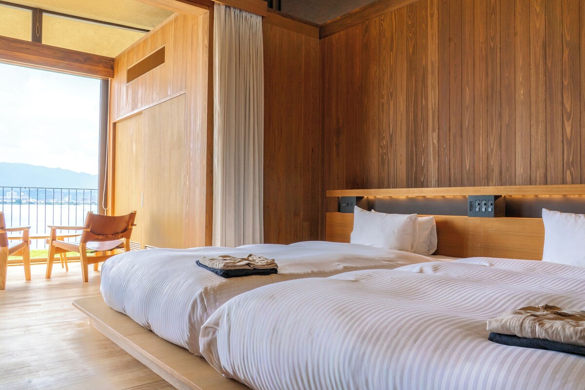 A hotel room with a large bed, a balcony with chairs, wooden paneling, and a view of the mountains.