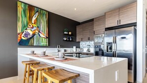Een moderne keuken met een centraal eiland, houten krukken, roestvrijstalen apparatuur en een groot abstract schilderij aan de muur.