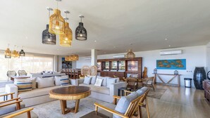 Een ruime zithoek met een grote bank, houten stoelen, een ronde salontafel en hangende rieten lampen.