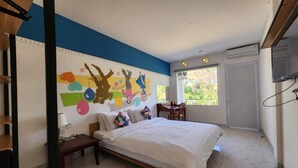 A bedroom with a bed, a desk, a chair, a window, and a wall with colorful artwork. - Stylish Double Suite with Balcony & Open Views (Bali)