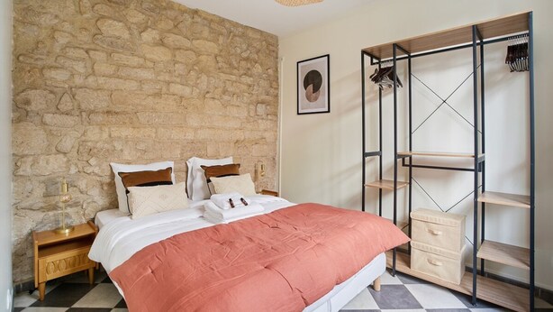 Quarto com parede de pedra, cama com manta rosa, mesa de cabeceira de madeira, guarda-roupa de metal e luminária pendente.