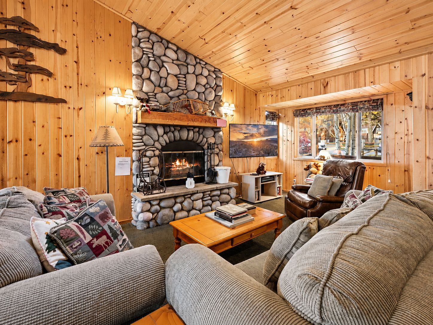 A cozy cabin interior with a stone fireplace, wooden walls, a leather sofa, and a television.