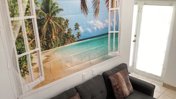 Ein Wohnzimmer mit großem Fenster, das eine tropische Strandmalerei zeigt, einem dunklen Sofa mit Kissen und einer zur Außenseite führenden Tür.