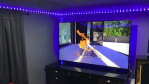 Ein Zimmer mit einem Flachbildfernseher auf einem Standfuß, einer roten Decke auf dem Boden und einem Fenster mit Blick auf Grünfläche.