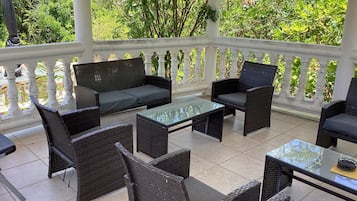Un espacio de estar al aire libre techado con mobiliario de mimbre y una mesa de vidrio, rodeado por una barandilla blanca.