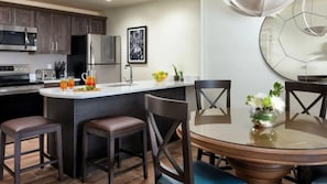 A modern kitchen with a central island, bar stools, a dining table with chairs, and a large clock on the wall.
