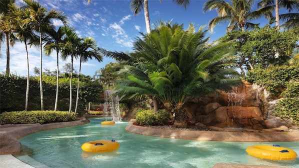 Un parc aquatique avec une piscine, des bouées gonflables, des palmiers et une cascade.