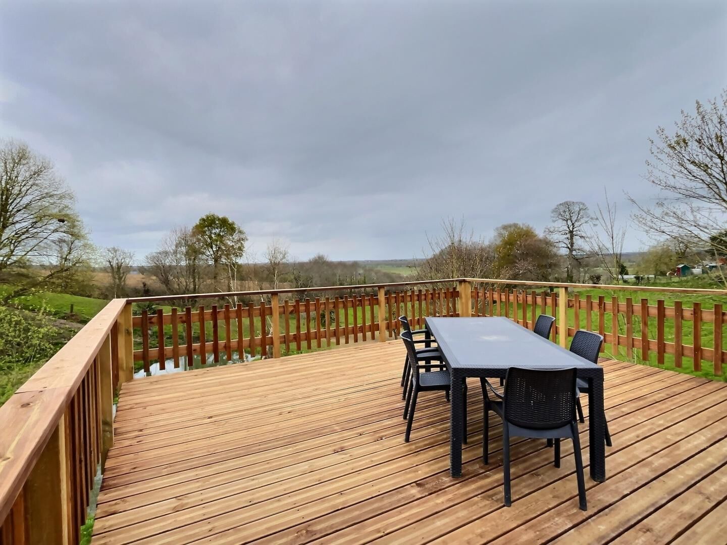 Eine Holzterrasse mit Tisch und Stühlen, umgeben von einem Holzzaun, mit Blick auf eine ländliche Landschaft.