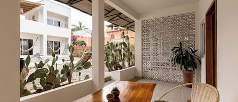 Une table en bois avec un coussin, une chaise en osier et une vue sur une cour intérieure agrémentée de cactus et d’un bâtiment.