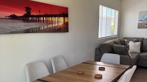 A dining area with a wooden table and chairs, a painting of a pier, a window with blinds, and a sofa in the background.