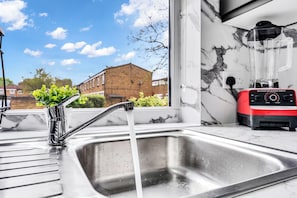 A kitchen with a stainless steel sink, a red and black blender, and a window with a view of a brick building.