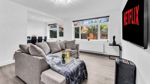 A modern living room with a grey sofa, a coffee table with a bottle and glass, and a large TV screen displaying 'NETFLIX'. - Ideal choice for those seeking comfort, convenience, and contemporary lifestyle (London)