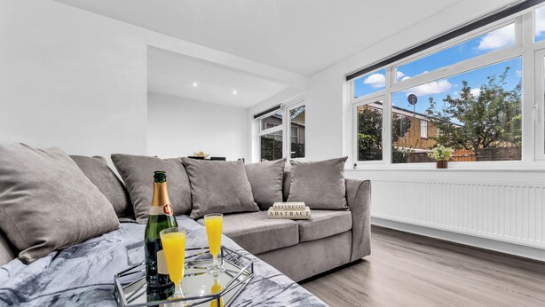 Een moderne woonkamer met een grijze bank, een glazen bijzettafeltje met een fles en glas, en uitzicht op het exterieur door grote ramen.