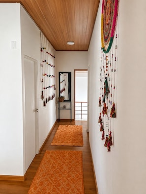 Ein Korridor mit Holzboden, weißen Wänden und einer Decke mit Holzpaneelen. Zwei orangefarbene Teppiche liegen auf dem Boden, an der Wand befindet sich ein Spiegel und links eine Tür.